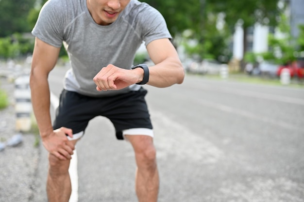 Atleta masculino asiático exhausto con las manos en la rodilla comprobando su frecuencia cardíaca en el reloj inteligente