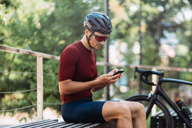 Atleta masculino caucásico que usa un teléfono inteligente moderno mientras se sienta en un banco cerca de una bicicleta deportiva negra Ciclista con ropa deportiva, casco de seguridad y anteojos espejados