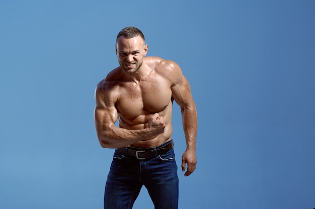 Atleta masculino com corpo musculoso mostra seu bíceps em estúdio, fundo azul. Um homem atlético, esportista sem camisa e calça jeans, estilo de vida ativo e saudável