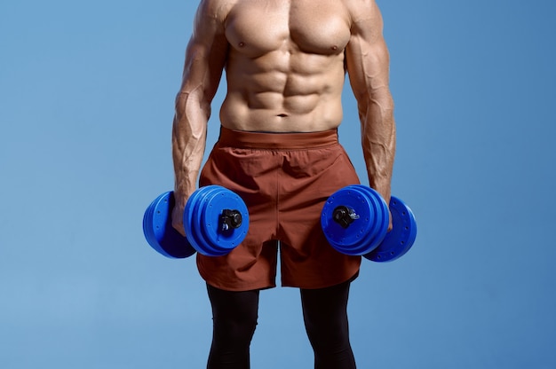 Atleta masculino con cuerpo musculoso sostiene pesas en estudio, fondo azul. Un hombre con complexión atlética, deportista sin camisa en ropa deportiva, estilo de vida activo y saludable