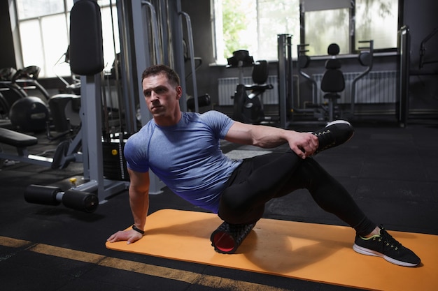 Atleta masculino enfocado usando rodillo de espuma estirando los músculos después de hacer ejercicio en el gimnasio