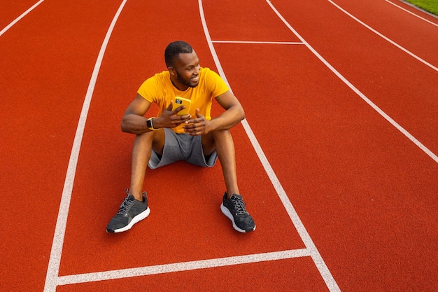 Foto atleta masculino feliz y atractivo sentado en la pista del estadio relajándose tomando un descanso después de correr