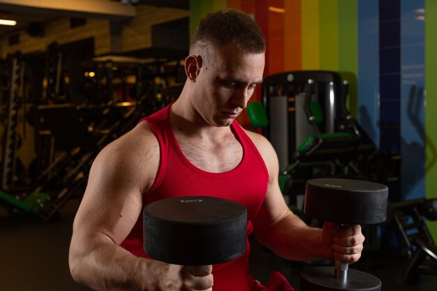 Un atleta masculino fuerte con una camiseta roja sostiene tensamente pesas y mira hacia abajo de pie en el gimnasio.