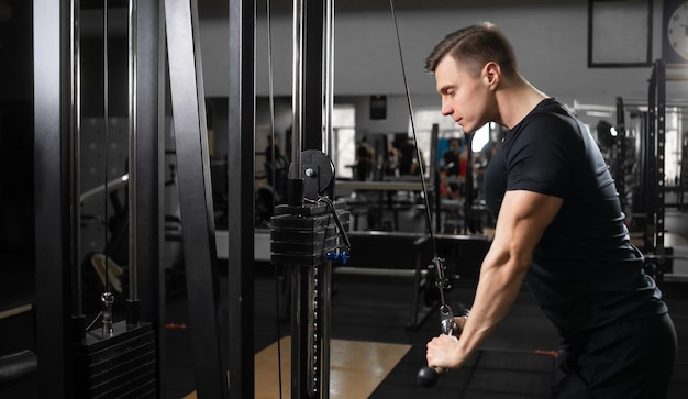 Un atleta masculino en el gimnasio se para detrás de una máquina de bíceps y tríceps y baja las barras de metal