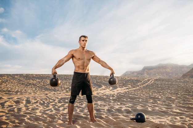 Atleta masculino haciendo ejercicios con dos pesas rusas en el desierto en un día soleado. Fuerte motivación en el deporte, entrenamiento de fuerza al aire libre