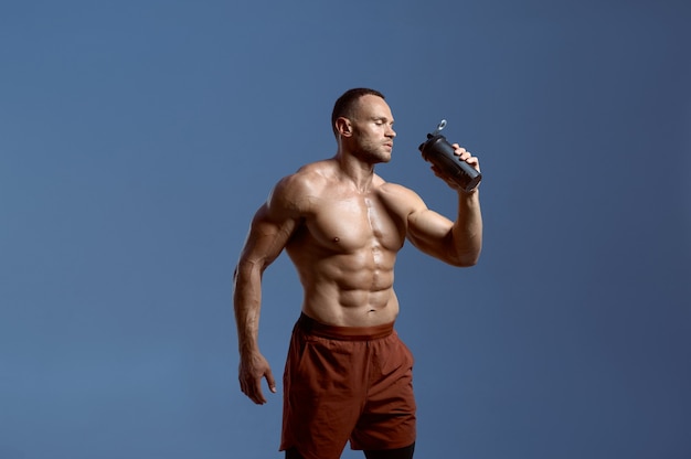 Atleta masculino musculoso bebe agua de botella deportiva, sesión de fotos en estudio, fondo azul. Un hombre con complexión atlética, deportista sin camisa en ropa deportiva, estilo de vida activo y saludable