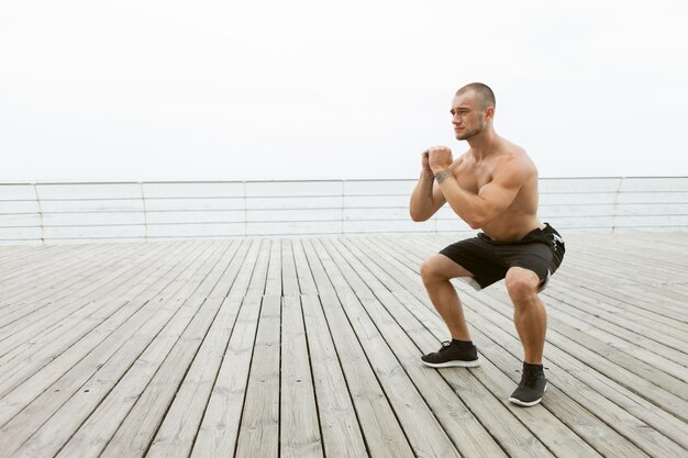 Atleta masculino musculoso com torso nu fazendo exercícios de agachamento na praia