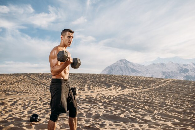 Atleta masculino musculoso fazendo exercícios com halteres no deserto em dia ensolarado. Forte motivação no esporte, treinamento de força ao ar livre, esportista com pesos