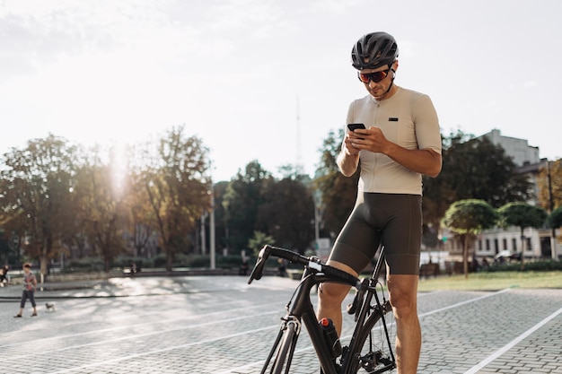 Atleta masculino no capacete e óculos sentado na bicicleta preta com smartphone moderno nas mãos Ciclista caucasiana mensagens de texto no celular enquanto fazia uma pausa durante o treino