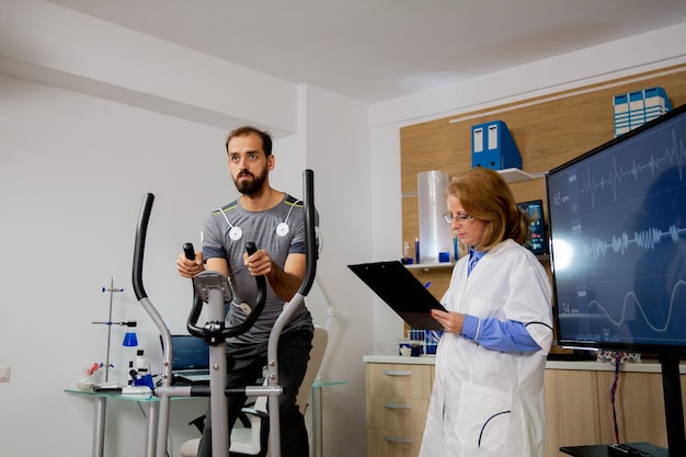 Atleta masculino que hace un esfuerzo físico en el paso a paso y tiene electrodos adheridos y el médico toma notas en el portapapeles.