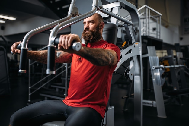 Atleta muscular na máquina de exercícios no ginásio. Homem barbudo fazendo exercícios no clube esportivo, estilo de vida saudável