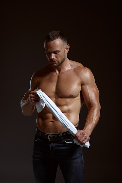 Atleta musculoso masculino sostiene su camisa en las manos en estudio, fondo oscuro. Un hombre con complexión atlética, deportista sin camisa en pantalones vaqueros, estilo de vida activo y saludable