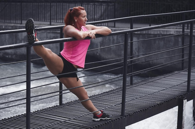 Atleta ruiva adulta de top rosa e shorts realiza exercícios de alongamento matinais no rio. estilo de vida saudável, ioga, fitness.