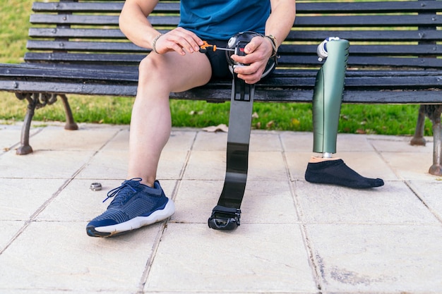 Foto atleta sentado en un banco en un parque ajustando una pierna protésica deportiva de fibra de carbono preparándose