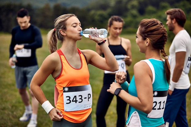 Atletas do sexo feminino se comunicando enquanto fazem uma pausa na maratona na natureza O foco está na mulher bebendo água de uma garrafa