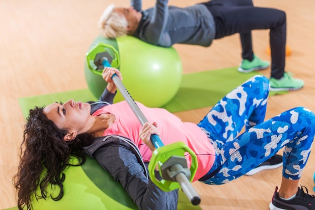 Atletas femeninas haciendo prensa de pecho con barra recostada sobre una pelota suiza durante el entrenamiento grupal en el gimnasio