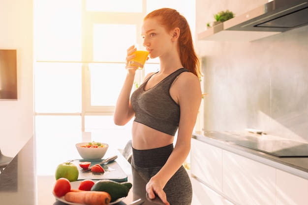 Atlética joven pelirroja en la cocina bebe un vaso de jugo de fruta centrifugado