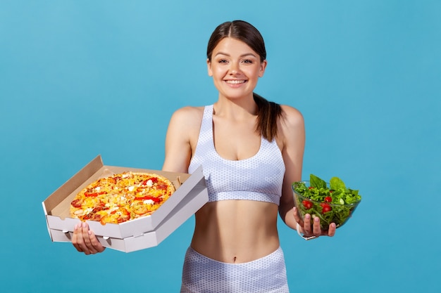 Atlética mulher segurando uma caixa de pizza e uma tigela com salada fresca e suculenta,