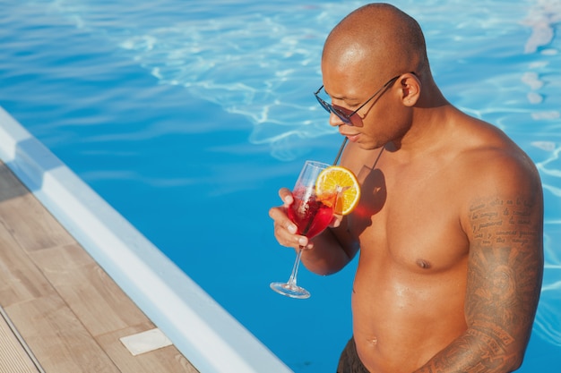 Atlético hombre africano tatuado sorbiendo su bebida, relajarse cerca de la piscina, copie el espacio. Joven disfrutando de sus vacaciones de verano, bebiendo cócteles en la piscina