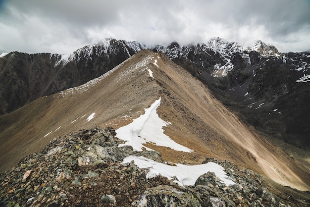 Atmosférico paisaje alpino minimalista hasta una enorme cadena montañosa nevada. Firn o nieve en combe rocky hill. Cielo de nubes sobre gran cresta nevada. Roca del arroyo Boulder. Majestuoso paisaje a gran altura