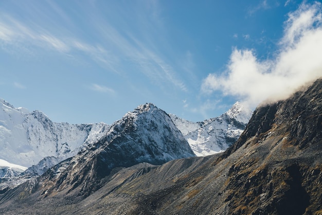 Atmosphärische Alpenlandschaft mit hohem schneebedecktem Berg mit spitzer Spitze unter Zirruswolken im Himmel. Großer schneebedeckter Berg im Sonnenschein. Niedrige Wolken auf schwarzen Felsen und spitzer, weißschneebedeckter Gipfel im Sonnenlicht