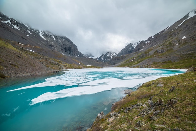 Atmosphärische Berglandschaft mit gefrorenem Alpensee und hohen schneebedeckten Bergen Ehrfürchtig bewölkte Landschaft mit eisigem Bergsee auf Hintergrund von Schneebergen in niedrigen Wolken Szenische Ansicht zum Eissee