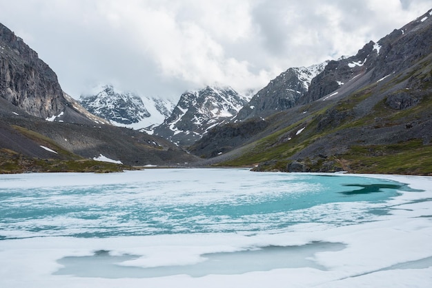 Atmosphärische Berglandschaft mit gefrorenem Alpensee und hohen schneebedeckten Bergen Ehrfürchtig bewölkte Landschaft mit eisigem Bergsee auf Hintergrund von Schneebergen in niedrigen Wolken Szenische Ansicht zum Eissee