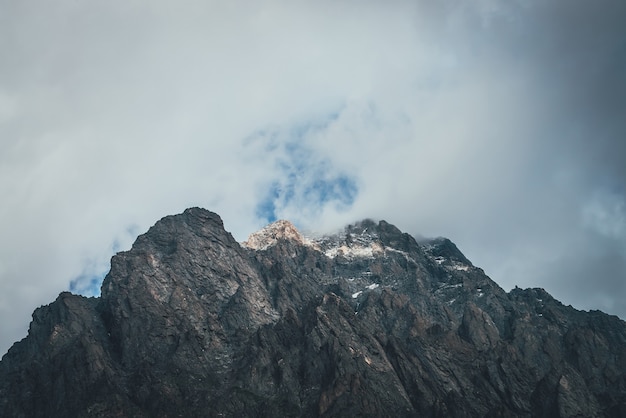 Atmosphärische Berglandschaft mit niedriger Wolke auf dem Berggipfel im Sonnenlicht. Dunkle Felsen mit Schnee und blauem Oberlicht im grauen bewölkten Himmel. Schöne Berglandschaft mit niedriger Wolke auf felsiger Spitze bei Sonnenschein