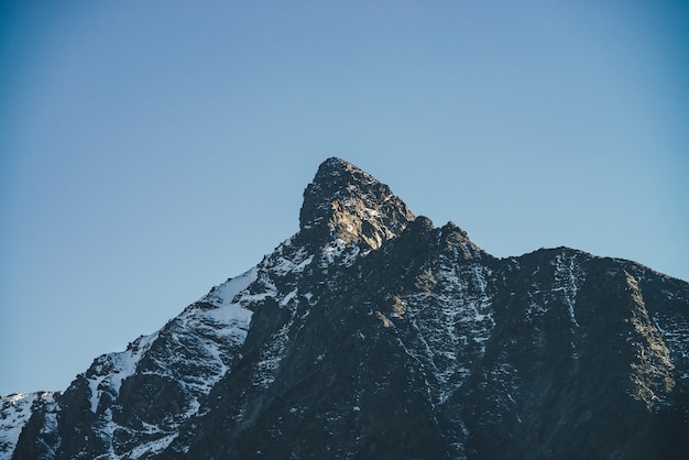 Atmosphärische Berglandschaft mit schwarzer Bergsilhouette mit scharfem Felsgipfel mit Schnee im Sonnenlicht. Alpenlandschaft mit dunklem schneebedecktem Berg mit spitzer Spitze bei Sonnenschein unter blauem Himmel