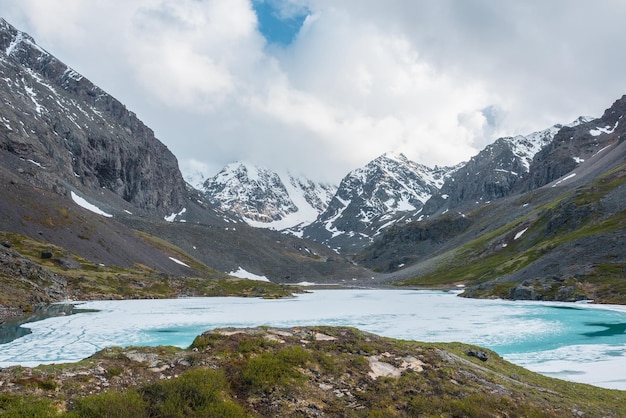 Atmosphärische Hochlandlandschaft mit gefrorenem Alpensee und hohen schneebedeckten Bergen Ehrfürchtige Landschaft mit eisigem Bergsee auf Hintergrund von großen Schneebergen in niedrigen Wolken Szenische Ansicht zum Eissee