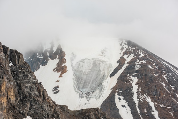 Atmosphärische Landschaft mit Gletscher auf felsigen Berggipfel in dichten niedrigen Wolken Ehrfürchtige Berglandschaft mit Gletscher zwischen scharfen Felsen in dicken niedrigen Wolken Hoher Berggipfel mit Gletschereis im Nebel