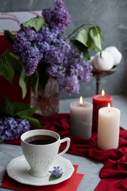 Atmosphärisches Foto: schwarzer Kaffee und eine weiße Tasse, ein Strauß Flieder, ein paar große Kerzen, Marshmallows auf grauer Oberfläche