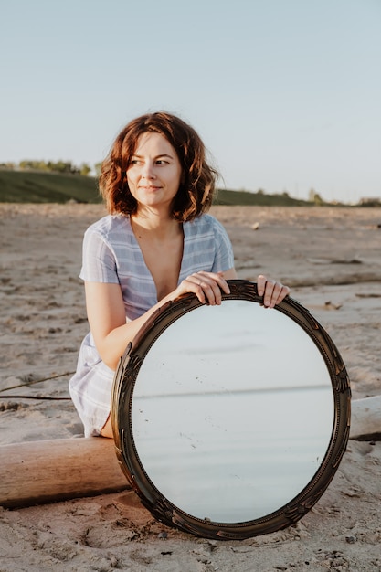 Atmosphärisches Lebensstilfoto im Freien der jungen schönen dunkelhaarigen Frau im blauen Kleid, das am Strand sitzt und Spiegel hält.