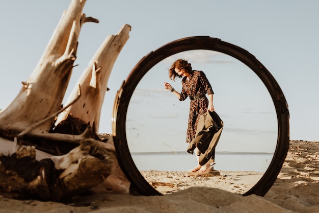 Atmosphärisches Lebensstilfoto im Freien der jungen schönen dunkelhaarigen Frau im Kleid und in der Jacke, die auf dem Strand im Spiegelbild spiegelten.