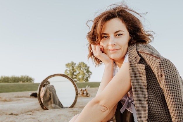 Atmosphärisches Lebensstilfoto im Freien der jungen schönen dunkelhaarigen Frau in den Jacken, die auf dem Strand und im Spiegelungsspiegel sitzen.