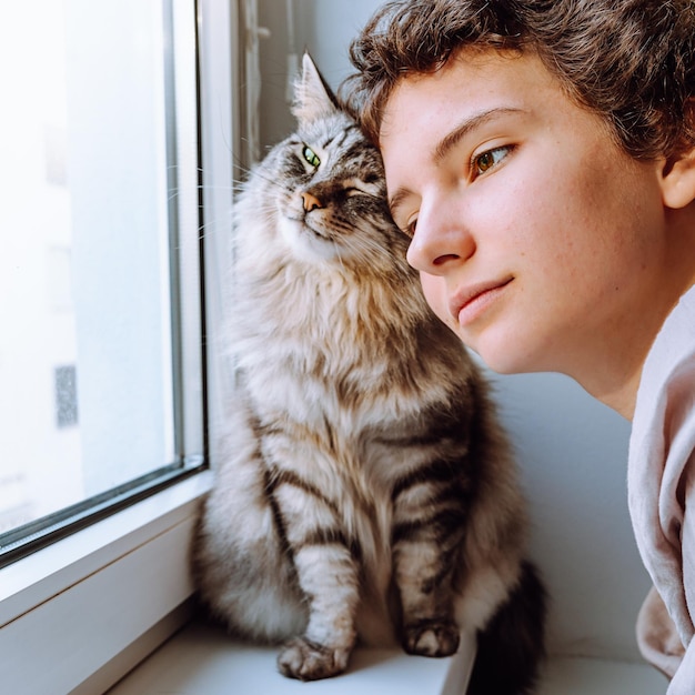 Atractiva adolescente junto al amado gato abrazándose mientras mira por la ventana