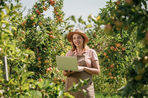 Atractiva agrónoma o agricultora con una computadora portátil parada en un huerto de manzanas y revisando la fruta toma notas Concepto de agricultura y jardinería Nutrición saludable