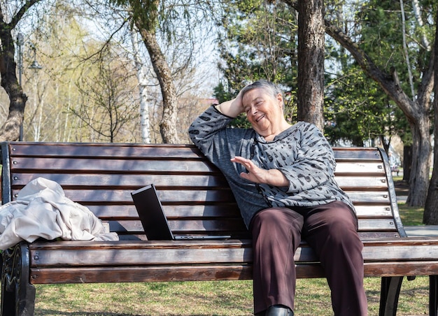 Atractiva anciana caucásica sentada en un banco en el parque usando una computadora para chat remoto en línea