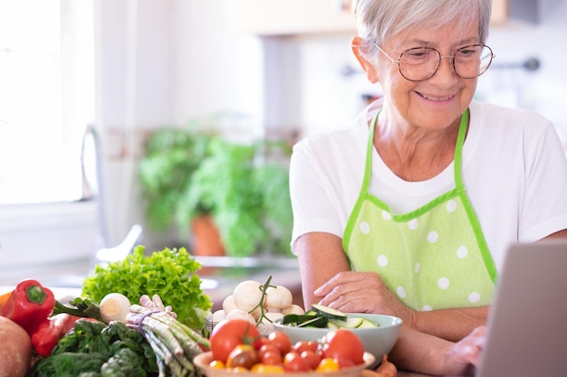 Atractiva anciana preparando verduras en la cocina de la casa navegando en la computadora portátil en busca de nuevas recetas Personas mayores caucásicas disfrutando de una alimentación vegetariana saludable