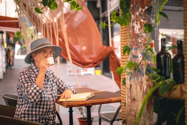 Atractiva anciana con sombrero sentada en la mesa de café leyendo un libro mientras bebe un café espresso dama caucásica disfrutando del tiempo libre y la jubilación