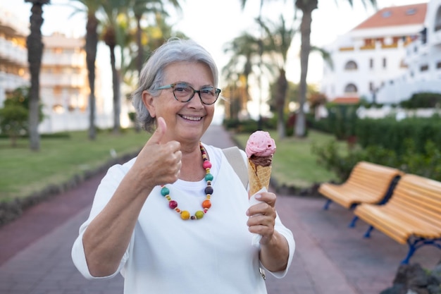 Atractiva anciana sonriente feliz disfrutando de un cono de helado levantando el pulgar Abuela caucásica mayor disfrutando de comida dulce y vacaciones en un lugar tropical mirando a la cámara