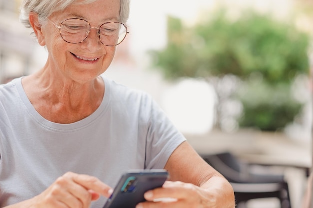 Atractiva anciana sonriente usando un teléfono móvil sentada en la mesa del bar señora madura escribiendo un mensaje en el teléfono celular