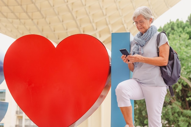 Atractiva anciana sonriente de vacaciones en Sevilla España parada cerca de un gran corazón rojo sosteniendo un teléfono móvil en la mano