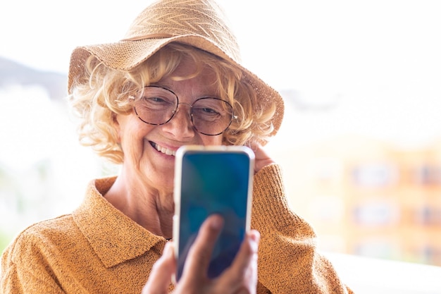 Atractiva anciana vestida de amarillo con sombrero y anteojos al aire libre en video chat con teléfono móvil anciana rubia rizada disfrutando de la tecnología y las redes sociales