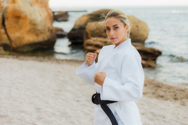 Atractiva artista marcial femenina en un kimono blanco con un cinturón negro se encuentra en una posición de combate en la playa salvaje