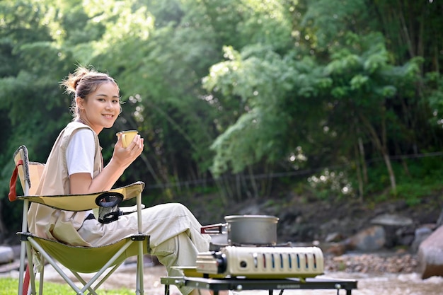 Atractiva campista asiática disfrutando del café de la mañana y la vista de la naturaleza cerca del río