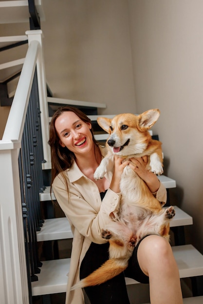 Atractiva chica abraza y juega con el perro corgi en casa Welsh Corgi Pembroke con su dueña mujer sentada en las escaleras