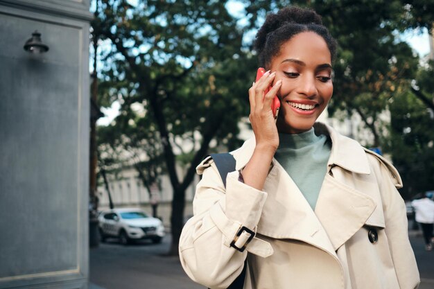 Atractiva chica afroamericana sonriente con elegante gabardina hablando alegremente por celular en la calle de la ciudad