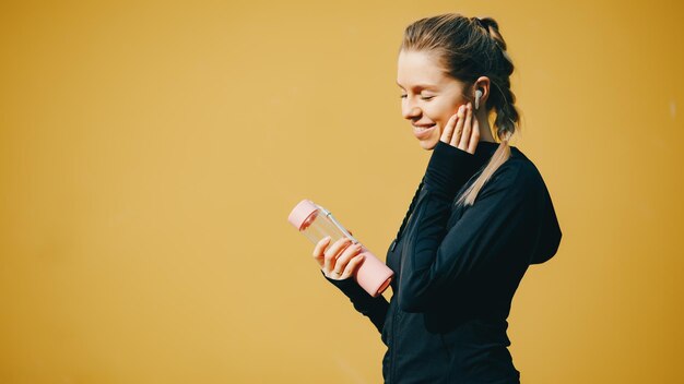 Atractiva chica caucásica sobre fondo amarillo aislado con auriculares inalámbricos en traje deportivo hacer ejercicio correr y beber agua