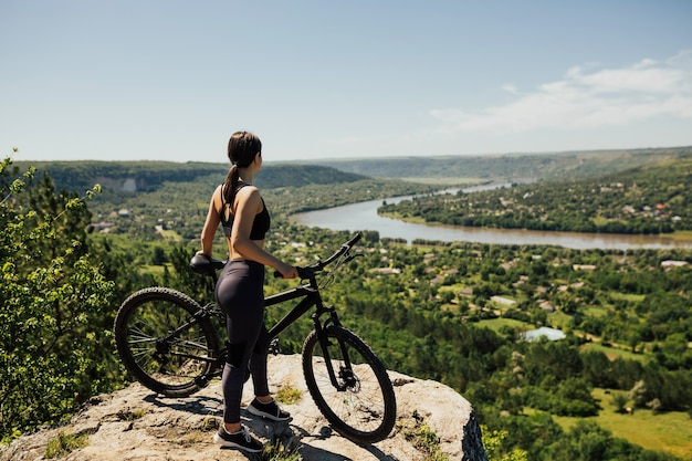 Foto atractiva chica deportiva de pie sobre la roca en la montaña con el telón de fondo del río, la ciudad y el cielo azul